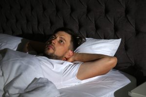 Man lying awake in bed at night, looking tired and unable to sleep, illustrating how summer heat and stress can disrupt rest during addiction recovery.